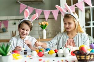 duas crianças felizes em orelhas de coelho pintam ovos de Páscoa na mesa. foto