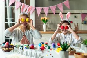 irmãzinha positiva e alegre e seu irmão, preparando-se para a Páscoa, sentados à mesa, segurando ovos coloridos e pintados no lugar dos olhos foto