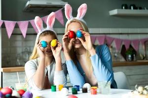 filha pequena, positiva e alegre e sua mãe, preparando-se para a Páscoa, sentadas à mesa, segurando ovos coloridos pintados no lugar dos olhos