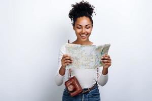 jovem bonita e alegre segurando um mapa e uma câmera em um fundo de parede branca foto