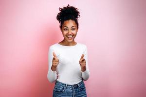 mulher alegre e confiante aponta o dedo indicador para a câmera, sorri amplamente, vestida com roupas da moda, modelos em uma parede rosa pastel foto