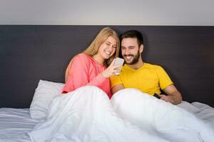 jovem doce casal dentro cama olhando às uma Móvel telefone foto