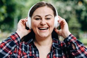 adorável senhora em pé na cidade e ouvindo a música favorita em fones de ouvido. retrato ao ar livre da sonhadora menina europeia usa fones de ouvido brancos. foto