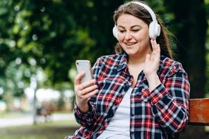 mulher linda e charmosa com protetores de ouvido, sentada em um banco, ouvindo algo em fones de ouvido, segurando um smartphone foto