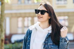garota atraente com cabelos escuros, vestida no estilo casual, com óculos escuros, andando pela rua. foto