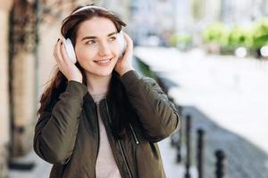 garota alegre e atraente caminha pela rua em fones de ouvido sem fio. ouve sua música favorita foto