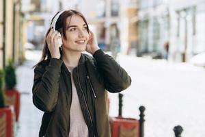 garota alegre e atraente caminha pela rua em fones de ouvido sem fio. ouve sua música favorita foto