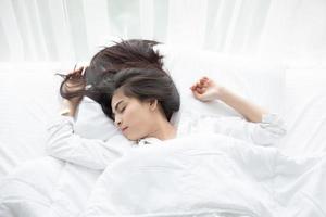mulher asiática linda jovem sorridente mulher sentada na cama e alongamento pela manhã no quarto, depois de acordar em sua cama totalmente descansada e abrir as cortinas pela manhã para tomar ar fresco. foto