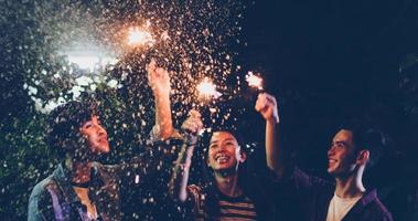 Grupo asiático de amigos fazendo churrasco ao ar livre no jardim, rindo com bebidas alcoólicas de cerveja e mostrando um grupo de amigos se divertindo com estrelinhas à noite, foco suave foto