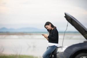 Mulher asiática usando telefone celular enquanto olha e homem estressado sentado após uma quebra de carro na rua foto