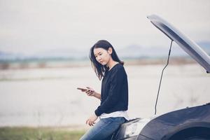 Mulher asiática usando telefone celular enquanto olha e homem estressado sentado após uma quebra de carro na rua foto