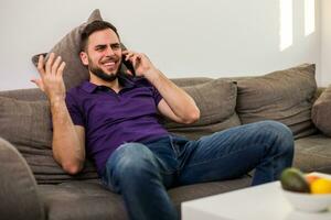 jovem homem homem usando Móvel telefone enquanto sentado em sofá dentro dele vivo sala. foto