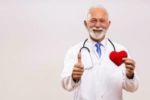 retrato do Senior médico com estetoscópio mostrando coração forma e polegar acima em cinzento fundo. foto