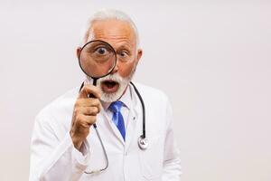 retrato do chocado Senior médico médico com lupa em cinzento fundo. foto