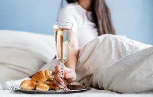 jovem morena sentada acordada na cama com balões em forma de coração vermelho e decorações bebendo champanhe comendo croissants