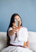 jovem morena sentada acordada na cama com balões em forma de coração vermelho e decorações bebendo champanhe comendo croissants