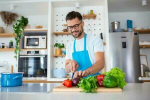 bonito jovem homem homem ficar de pé às moderno cozinha Picar legumes preparar fresco vegetal salada para jantar ou almoço, jovem masculino cozinhando às casa faço café da manhã Segue saudável dieta, vegetariano conceito foto