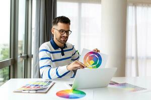 jovem freelance gráfico desenhador cara trabalhando com computador portátil e cor amostras, sentado às escrivaninha dentro casa escritório, escolhendo cor gama para Novo Projeto projeto, lado Visão com cópia de espaço foto