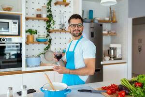 casa cozinhar. solteiro homem dentro avental olhando às a janela, bebendo vinho a partir de uma vidro enquanto cozinhando dentro a cozinha, cozinhando às casa foto