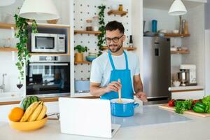 uma jovem homem gasta uma dia às lar, prepara Comida do legumes dentro a cozinha, uma homem dentro casa roupas e com uma barba, usa uma computador portátil para aprender conectados cozinhando foto