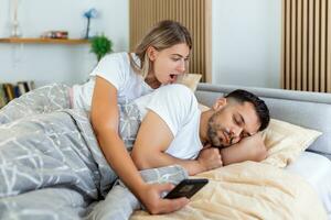 mulher é ciumenta e desconfiada e espiona o smartphone do parceiro enquanto ele dorme no quarto. a esposa está espionando o telefone do marido enquanto ele dorme. o conceito de desconfiança, ciúme foto