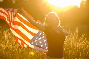 lindo jovem menina segurando a americano bandeira dentro a vento dentro uma campo do centeio. verão panorama contra a azul céu. horizontal orientação. foto