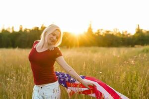 lindo jovem menina segurando a americano bandeira dentro a vento dentro uma campo do centeio. verão panorama contra a azul céu. horizontal orientação. foto