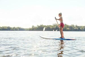 jovem atraente mulher em ficar de pé acima remo borda dentro a lago, sup foto