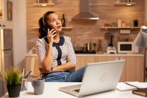 freelance mulher tendo uma telefone cobertura com uma cliente atrasado às noite a partir de casa cozinha. empregado usando moderno tecnologia às meia noite fazendo hora extra para trabalho, negócios, carreira, rede, estilo de vida. foto