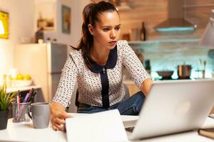 mulher verificação notas em prancheta enquanto trabalhando em computador portátil atrasado às noite dentro casa cozinha sentado às escrivaninha. empregado usando moderno tecnologia às meia noite fazendo hora extra para trabalho, negócios, carreira, rede. foto