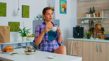 senhora desfrutando uma quente verde chá bebericando a partir de porcelana copo. mulher bebendo em pé às a mesa dentro a cozinha e olhando através a vidro segurando chá copo com natural, aromático ervas bebida. foto