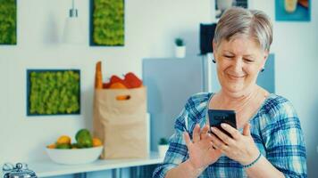 relaxado idosos mulher navegando em telefone dentro cozinha durante café da manhã. autêntico idosos pessoa usando moderno Smartphone Internet tecnologia. conectados comunicação conectado para a mundo, Senior lazer Tempo com gadget às aposentadoria era foto