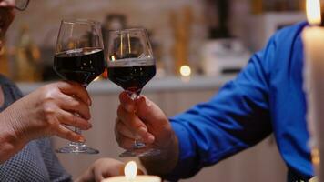 fechar acima do tilintar vermelho vinho óculos durante romântico jantar. feliz alegre Senior idosos casal jantar juntos dentro a acolhedor cozinha, desfrutando a refeição, a comemorar seus aniversário. foto