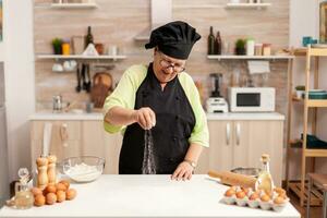 Senior senhora chefe de cozinha sorridente enquanto preparando pizza aspersão farinha em cozinha mesa. feliz idosos chefe de cozinha com uniforme aspersão, peneiramento peneirar cru ingredientes de mão. foto