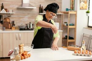 idosos mulher usando farinha para faço delicioso biscoitos em casa cozinha mesa espalhando farinha. feliz idosos chefe de cozinha com uniforme aspersão, peneiramento peneirar cru ingredientes de mão. foto