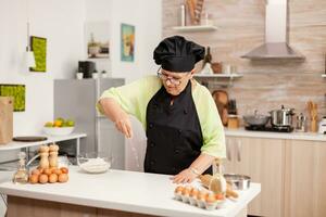 mulher é peneirar farinha em de madeira mesa dentro moderno cozinha. feliz idosos chefe de cozinha com uniforme aspersão, peneiramento peneirar cru ingredientes de mão cozimento caseiro pizza. foto