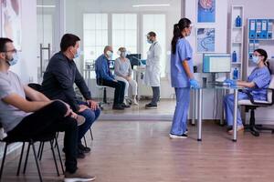 médico assistentes falando dentro hospital recepção vestindo proteção máscaras contra cobiçado 19 vírus enquanto pacientes esperando para exame. médico consultando pessoa dentro fundo durante global pandemia foto