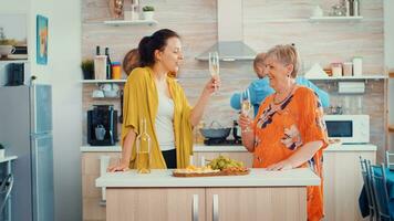 mãe e filha tilintar óculos do vinho sentado dentro a cozinha. estendido família a comemorar dentro jantar quarto bebendo uma vidro do vinho enquanto homens estão cozinhando dentro a fundo foto