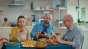 idosos pai assistindo fotos em Smartphone do jovem homem enquanto sentado dentro cozinha durante família jantar. multi geração, dois casais falando e comendo durante uma gourmet macho, desfrutando Tempo às casa