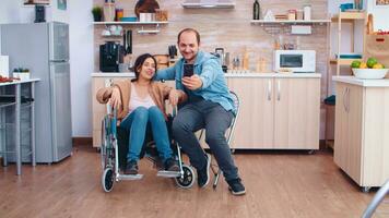 positivo mulher dentro cadeira de rodas e marido levando uma selfie dentro cozinha usando Smartphone. esperançoso marido com deficiente Desativado incapacidade inválido paralisia desvantagem pessoa Próximo para ele, ajudando dela para reintegrar foto