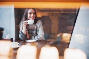 mulher jovem e bonita tomando café cappuccino com espuma perto da janela em um café foto