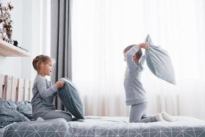 menino e menina encenaram uma luta de travesseiros na cama no quarto. crianças travessas batem umas nas outras nos travesseiros. eles gostam desse tipo de jogo foto