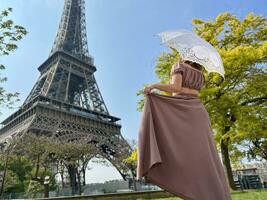 a partir de a costas nós Vejo uma menina dentro uma lindo grandes Castanho vestir dentro uma retro estilo com a guarda-chuva quem anda em para a eiffel torre segurando a vestir com 1 mão foto