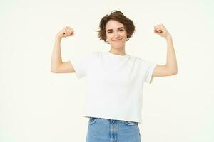 retrato do confiante e Forte jovem mulher, flexão bíceps, mostra dela músculos em braços com orgulhoso sorridente face, posando sobre branco fundo foto