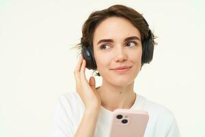 retrato do sincero jovem mulher, escuta música dentro sem fio fones de ouvido, segurando Smartphone, assistindo videos em Móvel telefone, em pé sobre branco fundo foto