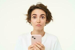 retrato do confuso menina em pé com Smartphone, levantando dela sobrancelhas dentro surpresa, em pé sobre branco fundo foto