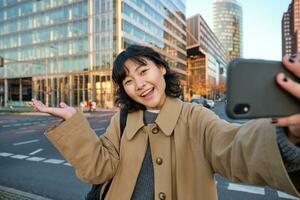 otimista ásia menina leva selfie com Smartphone dentro cidade Centro, mostrando alguma coisa atrás dela com sorridente face. turista vai passeios turísticos foto