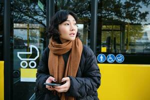 retrato do coreano menina olhando para dela ônibus em uma parar, segurando Móvel telefone, verificação agendar, Tempo mesa em Smartphone aplicativo foto