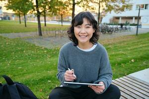 jovem garota, coreano artista ou arte aluna senta dentro parque com digital tábua, desenha com gráfico caneta, arranhões uma Projeto ou projeto, parece por aí foto
