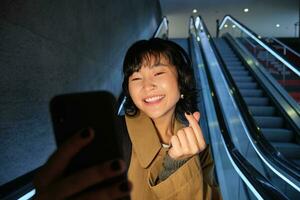 à moda sorridente ásia garota, levando selfie em Móvel telefone enquanto equitação escada rolante, indo baixa para metro estação, mostrando dedo coração e posando para foto